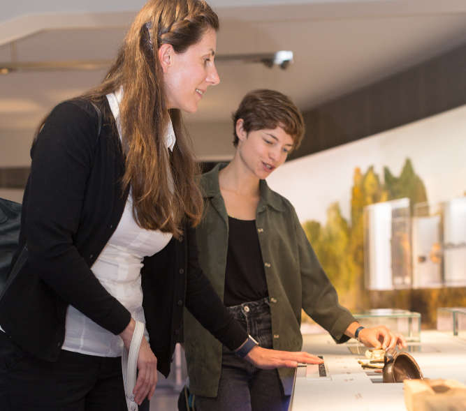 Blinde Museumsbesucherin tastet Braillebeschriftung eines Ausstellungsobjektes. Ihre Assistentin begleitet sie.