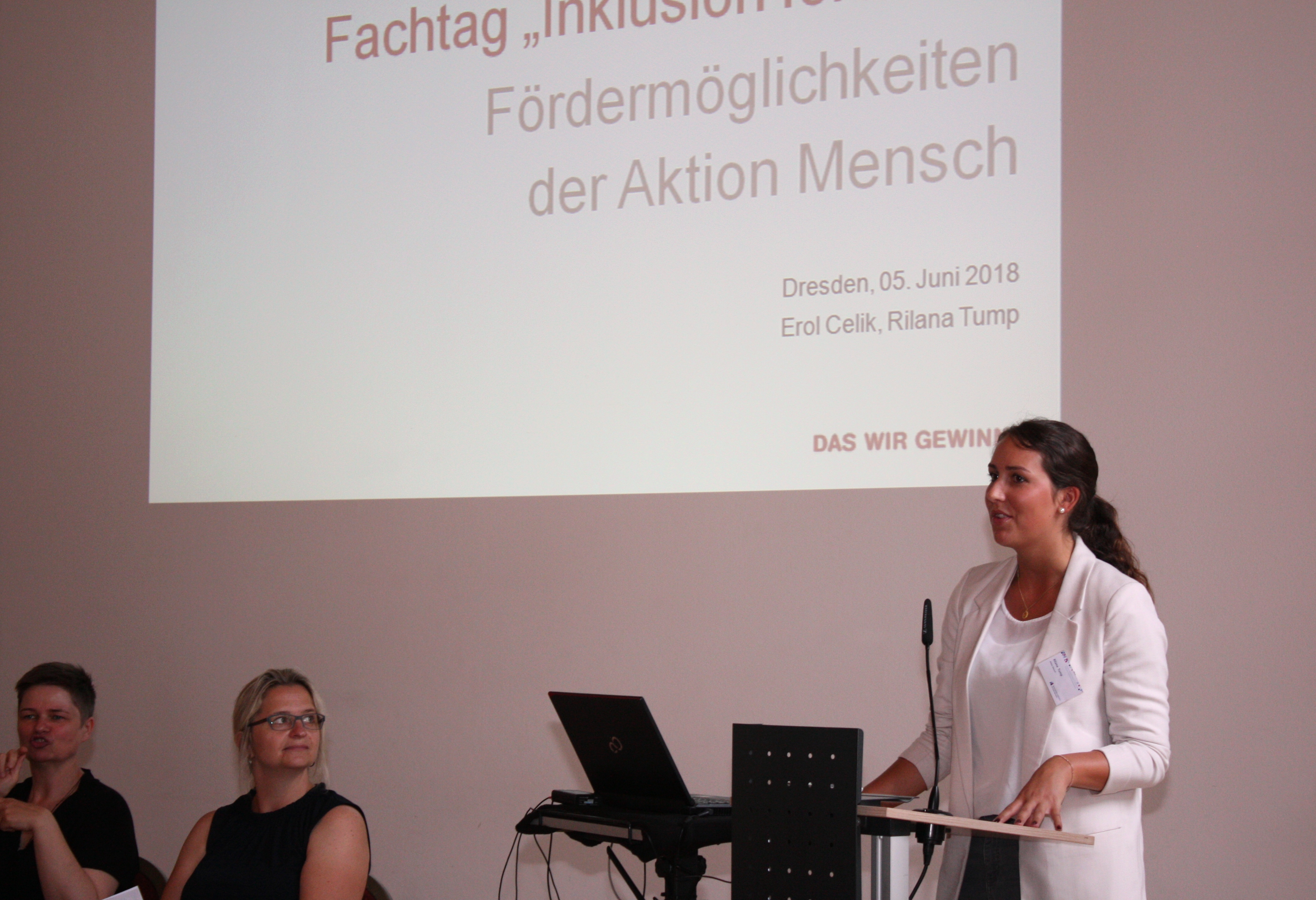 Referentin am Pult stehend, im Hintergrund Präsentation, links zwei Gebärdensprachdolmetscherinnen