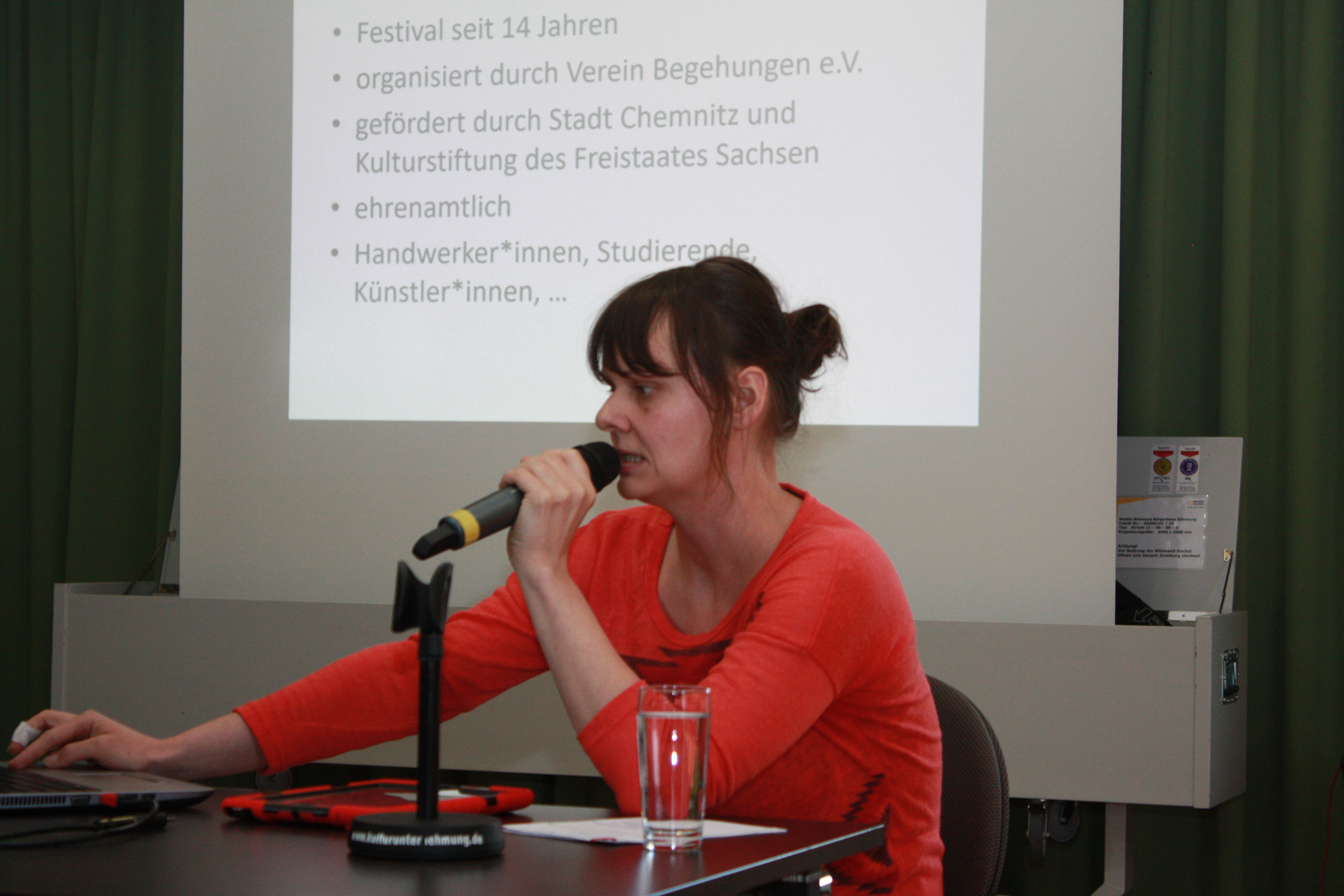 Eine Referentin sitzend vor Präsentation. Spricht ins Mikrofon.