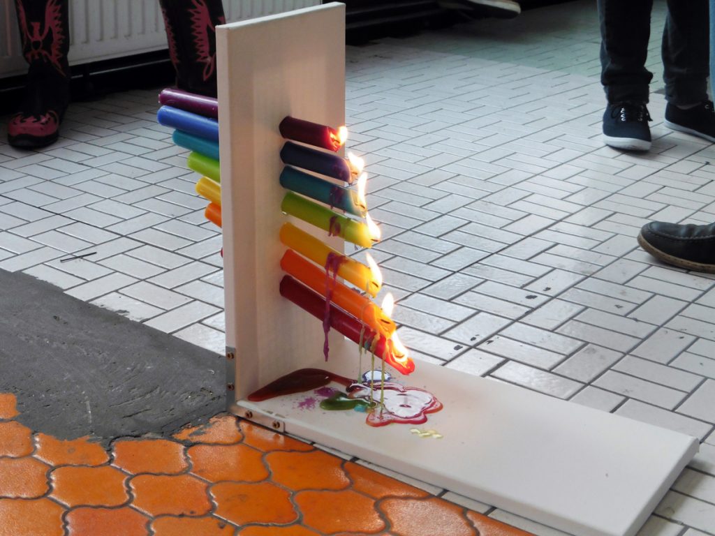 Installation „PRISMA“: Sieben farbige Kerzen stecken waagerecht in einer Leinwand, die auf dem Boden steht. Die Kerzen haben die Farben des Regenbogens, sind treppenförmig angeordnet und brennen. Der geschmolzene Wachs tropft von oben nach unten und vermischt sich dabei.