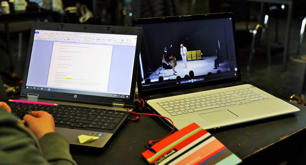 Auf einen Tisch stehen zwei Laptops nebeneinander. Sie sind aufgeklappt. der rechte Laptop zeigt ein Video, der linke einen Text. Zwischen den Laptops liegt ein Notizbuch.