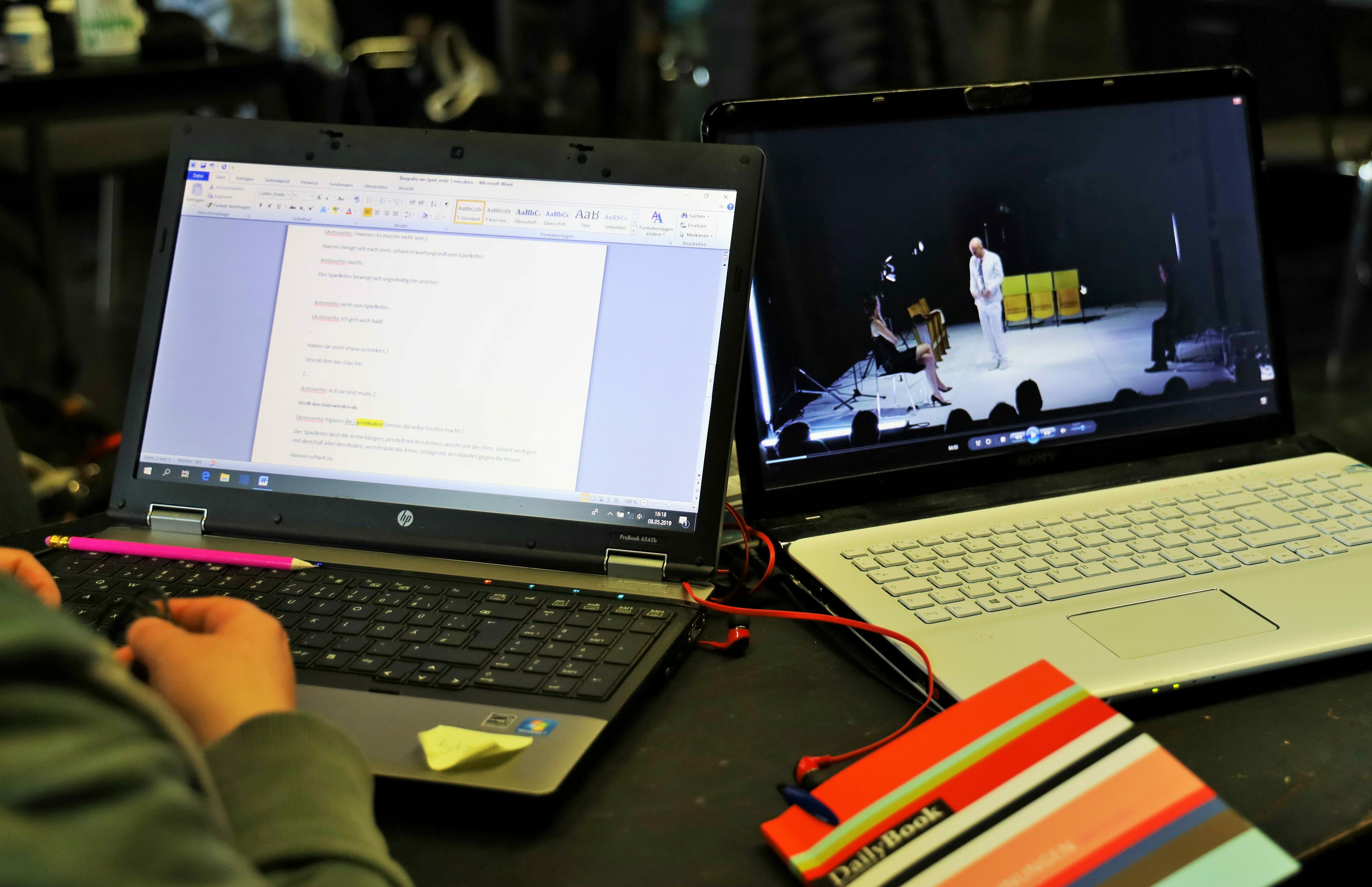 Das Foto zeigt zwei Laptops nebeneinander. Sie sind aufgeklappt. Der rechte Laptop zeigt ein Video, der linke einen Text. Zwischen den Laptops liegt ein Notizbuch.