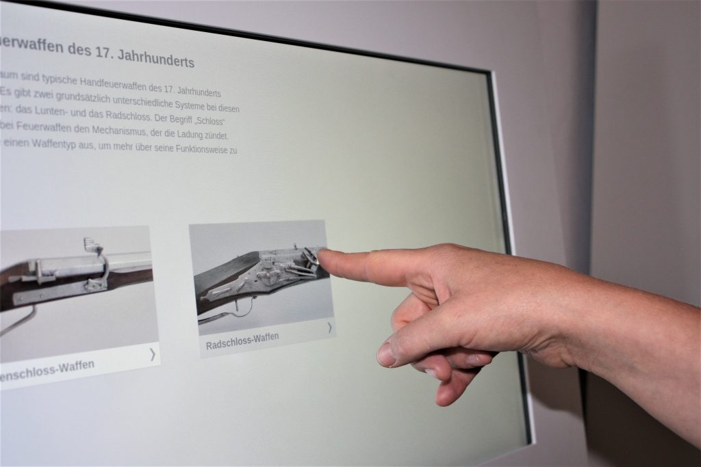 Zu sehen ist ein Touch Screen. Es werden Informationen und Bilder angezeigt. Ein Finger tippt auf das Bild.