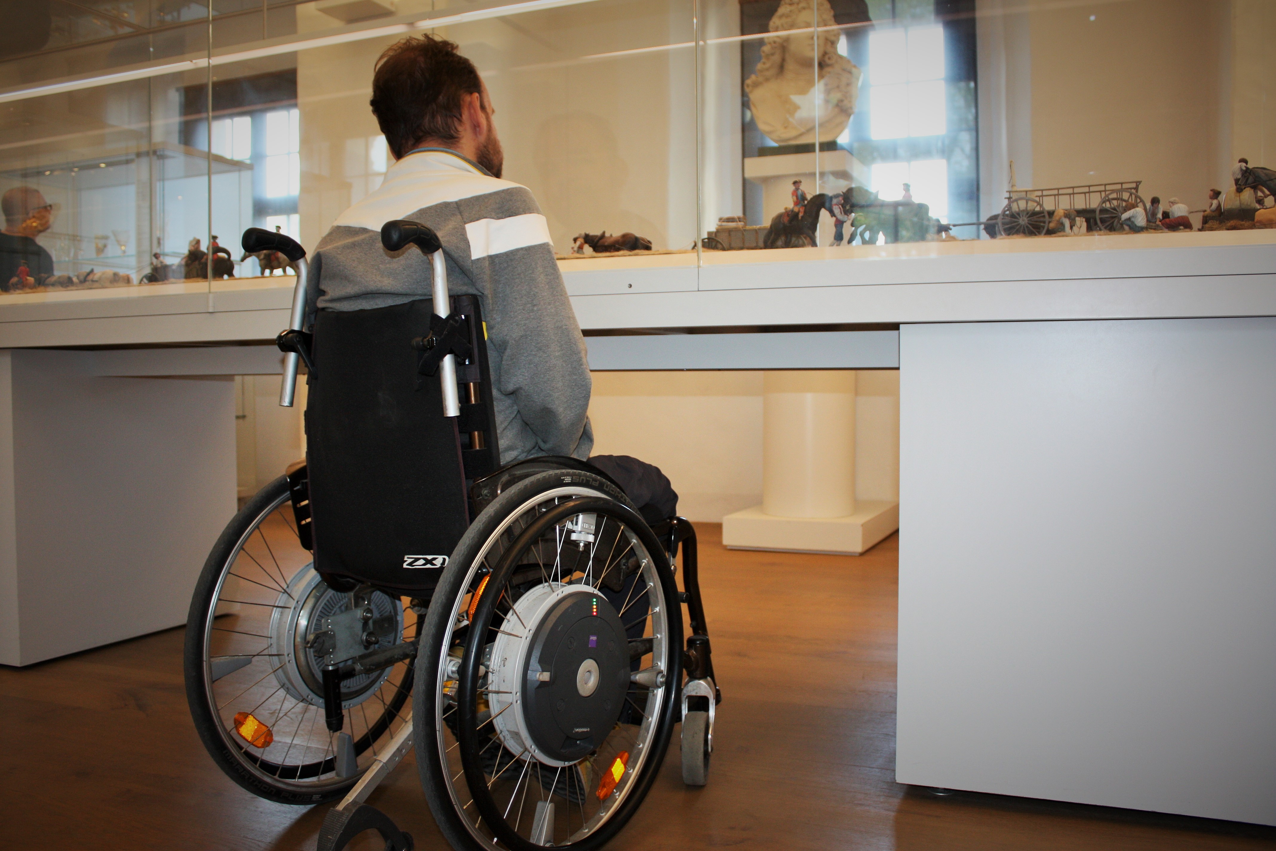 Ein Rollstuhlfahrer steht vor einer unterfahrbaren Ausstellungsvitrine. Er ist von hinten fotografiert.
