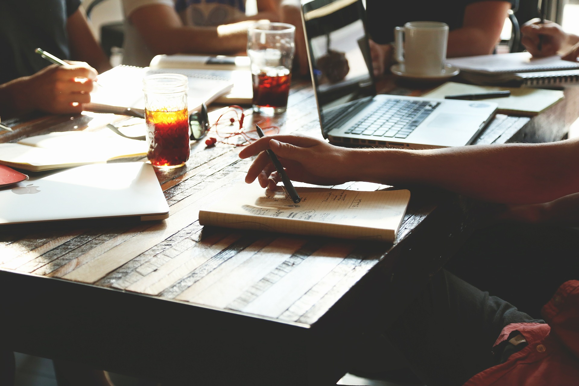 An einem Tisch sitzen sich vier Personen gegenüber. Von den Personen, die am Tisch sitzen, sind nur die Arme zu sehen. Die Personen halten einen Stift in der Hand. Auf dem Tisch liegen Notizblätter, ein Laptop, Kaffeetasse und zwei Gläser mit brauner Flüssigkeit.