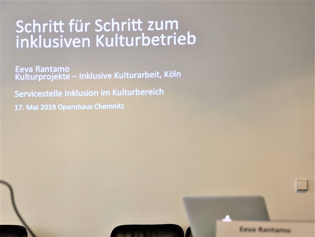 Das Foto zeigt eine Projektion der Präsentationsfolie des Workshops. Auf der Präsentationsfolie ist zu lesen: Schritt für Schritt zum inklusiven Kulturbetrieb.