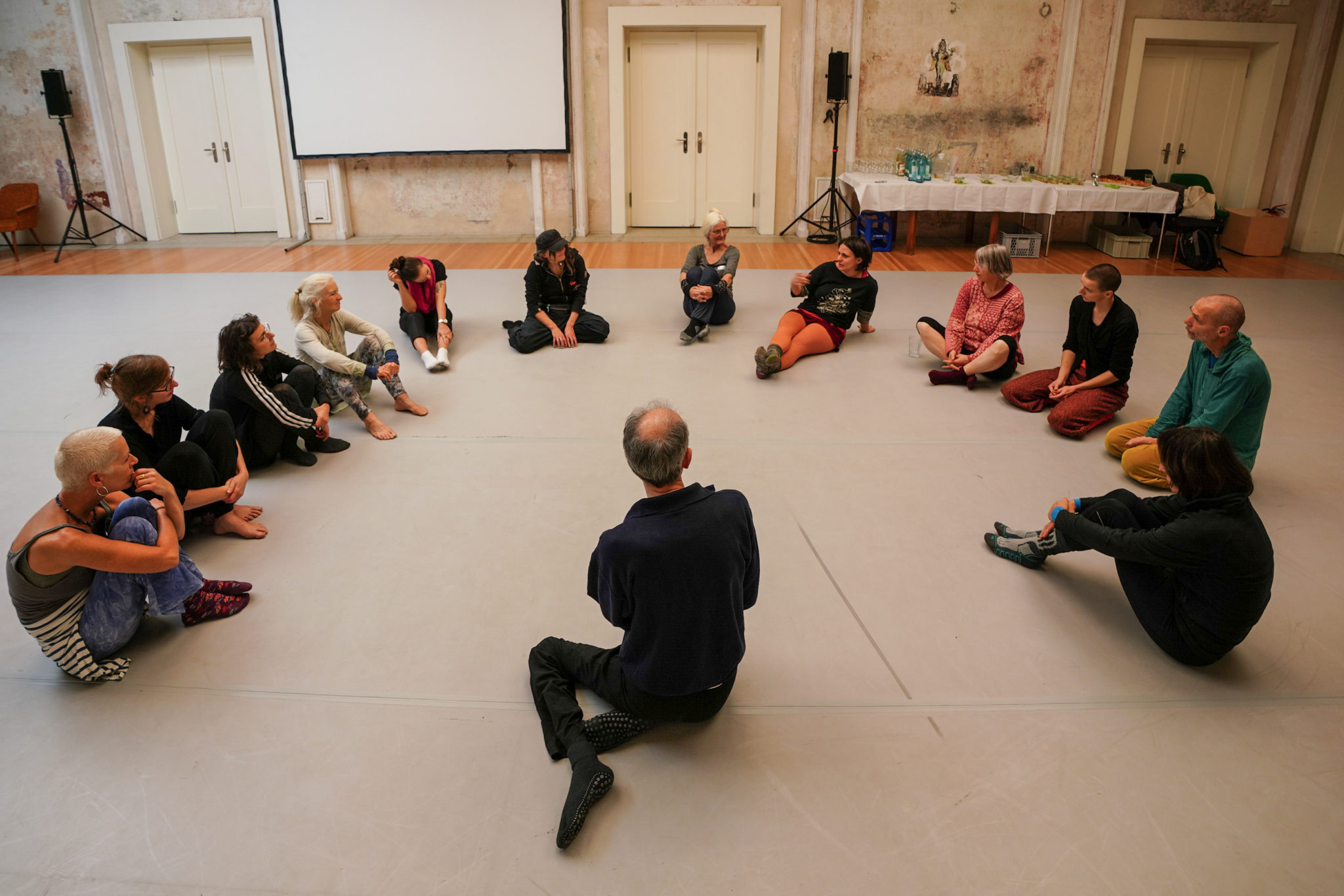 Das Foto zeigt Teilnehmende eines Workshops in einem Halbkreis sitzend auf dem Tanzboden. Der Kursleiter sitz vor ihnen. Auf dem Bild ist sein Rücken zu sehen.