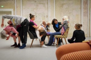 Foto, beim Fachtag Divers! am 18.12.2019 aufgenommen. Eine Gruppe führt den Tagungsteilnehmern ein Theaterstück vor. Es sind 9 Frauen, zum Teil Seniorinnen. Drei sitzen auf Sesseln. drei weitere umarmen jeweils eine sitzende Frau, beziehungsweise ien liegt auf dem Schoss einer sitzenden. Links stehen zwei gebeugte, ineinander verhakte Frauen.