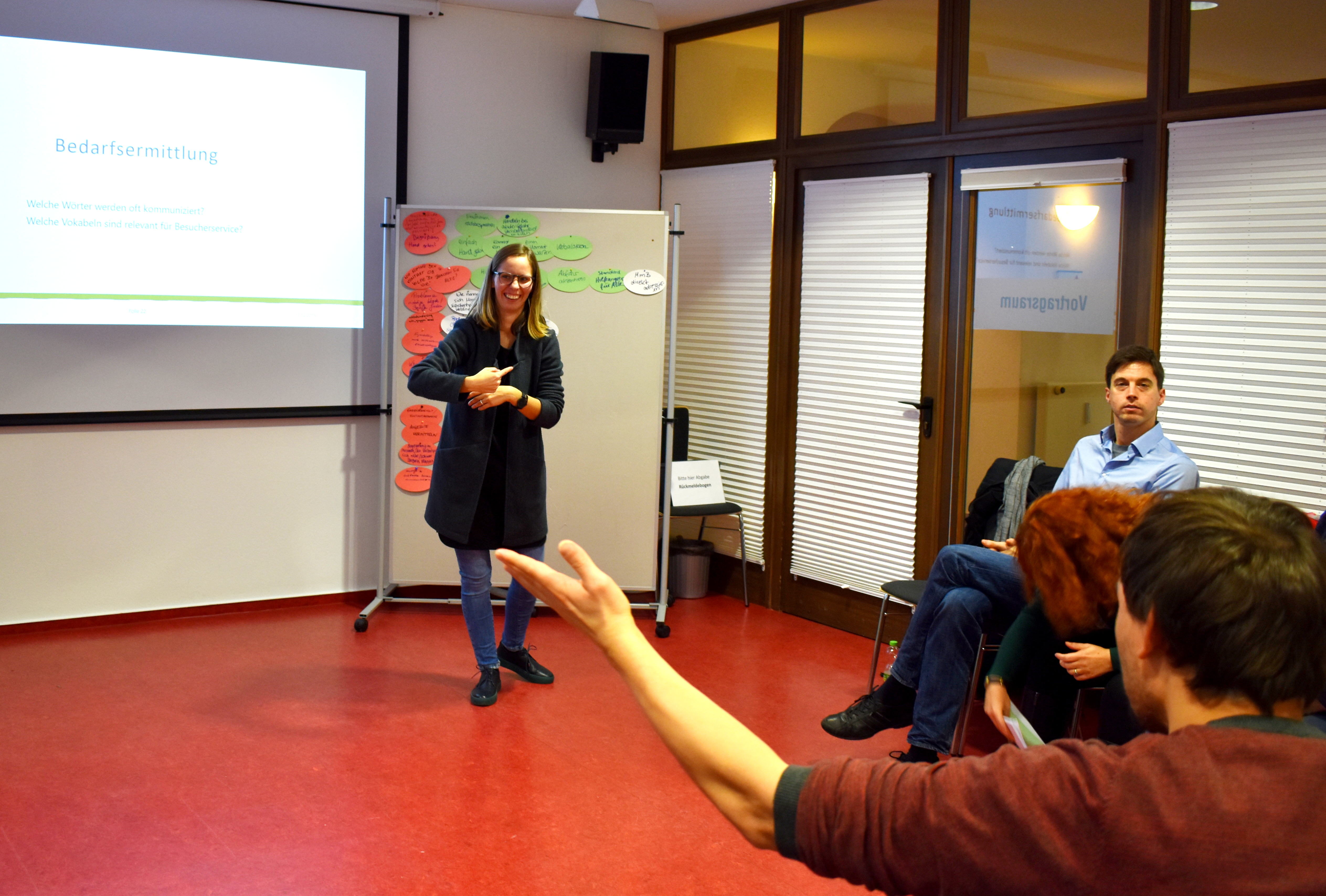 Das Foto zeigt eine Frau. Sie steht vor einer Pinwand referiert in Gebärdensprache. Auf der linken Seite befindet sich eine Leinwand an der Wand, auf die eine Powerpoint-Präsentation zu sehen ist. Vorn rechts ist ein Gebärdensprachdolmetscher während des Übersetzens zu sehen.