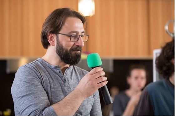 Auf dem Foto ist Düzgün Polat leicht von der Seite als Brustbild zu sehen. Er hält ein Mikro und spricht. Im Hintergrund befindet sich ein Saal und sitzende Zuhörer.