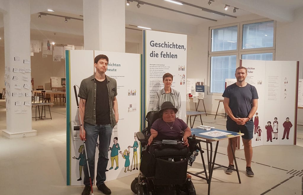 Das Foto zeigt vier Personen in einem Ausstellungsraum: links einen mann mit einen Langstock, rechts daneben eine Frau im Rollstuhl, dahinter eine Frau, rechts daneben einen Mann. Im Hintergrund sind Aufsteller und Ausstellungsobjekte zu sehen.