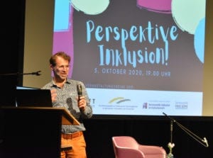 Zu sehen ist ein Ausschnitt der Bühne im LOFFT-DAS THEATER. Im Hintergrund befindet sich eine Leinwand auf die der Titel der Veranstaltung Perspektive Inklusion und die Kooperationspartner projeziert sind. Gustavo Fijalkow steht auf der linken Seite des Bildes an einem Pult und redet mit der einen Hand gestikulierend und mit der anderen Hand ein Mikrofon haltend. Hinter ihm ist ein leere Stuhl für die sich anschließende Podiumsdiskussion zu sehen.