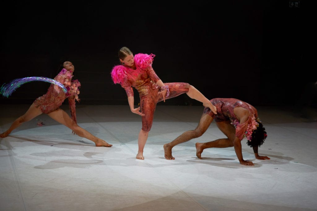 Szenenaufnahme der Forward Dance Companiy aus dem Stück JOY. Drei Tänzerinnen in rotbrauen Bodies auf der Bühne. Hellgrauer Bühnenboden, schwarzer Hintergrund. Lichtkegel über jeder Tänzerin. Die Tänzerin links steht im leichten Spagat nach vorn gebeugt, den Kopf nach hinten gelegt. Die tänzerin in der Mitte steht auf dem rechten Bein, das linke etwas angewinkelt, die Arme nach unten genommen und die Hände zueinander gedreht. Der Blick geht zur Tänzerin rechts, die auf allen vieren aussieht, als ob sie in den Startblöcken eines Sprints steht.