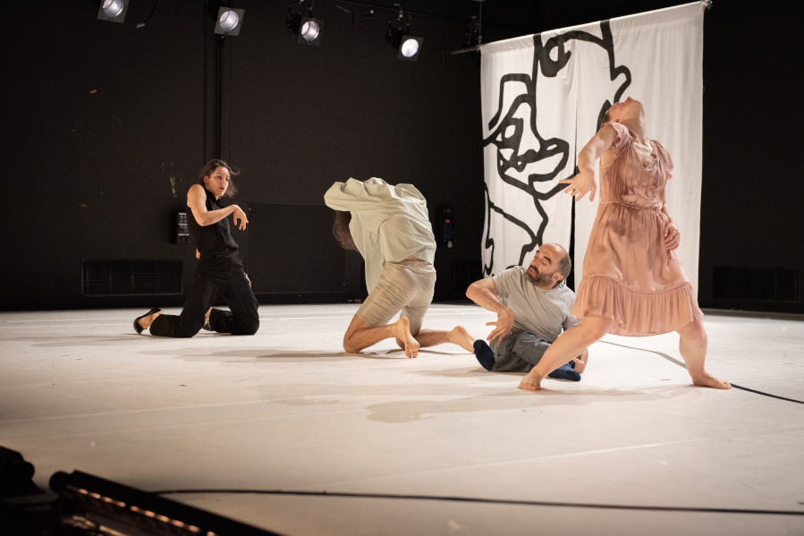 Szenenaufnahme der Forward Dance Companiy aus dem Stück WIR. Aufnahme von der rechten Seite auf die Bühne. Hellgrauer Bühnenboden, schwarzer Hintergrund vor dem ein großes weißes Textil hängt, auf dem eine schwarze Grafik ein abstrahiertes Gesicht zeigt. Auf der Bühne vier Tänzerinnen. Die Tänzerin links ist schwarz gekleidet und kniet zu den anderen Tänzerinnen gewendet. Ihr gegenüber eine weitere knieende Tänzerin in weiß. Als drittes ein kleiner Tänzer auf den Boden in grauer Kleidung und als vierte eine stehende Tänzerin im hellrosa Kleid von allen anderen abgewendet, leicht im Schritt, in die Knie gehend und den Kopf nach hinten werfend.