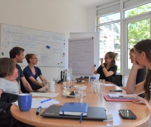 Fotografiert wurde ein Beratungsgespräch an einem langem Tisch. Vorn die kurze Seite des Tisches mit Laptop, Arbeitsbuch, Telefon und Stift. Auf der rechten Seite am Tisch drei Personen sitzend, die hinterste gebärdend. Auf der linken Seite ebenfalls drei Personen sitzend. Die vorderste im Rollstuhl sitzend. Der Tisch ist mit Arbeitsmaterialien und Getränken bestückt. Im Hintergrund beschriftete Tafeln und Blätter.