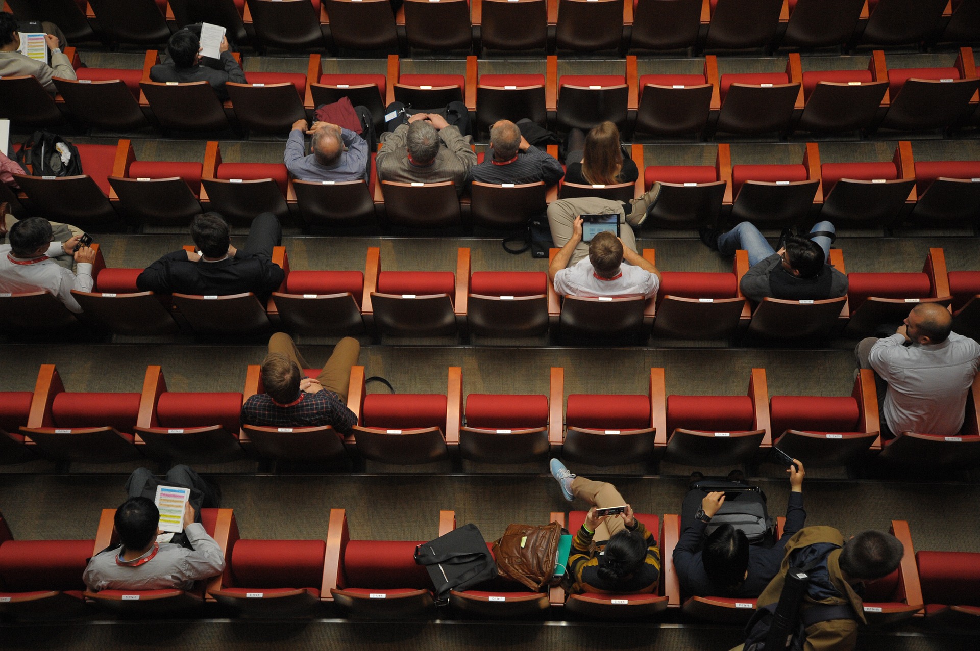 Das Foto zeigt ein Auditorium von oben. Das Auditorium ist mit roten Samtstuhlreihen bestuhlt. Die Stühle sind lückenhaft besetzt. Einige Personen haben einen Laptop auf dem Schoß.