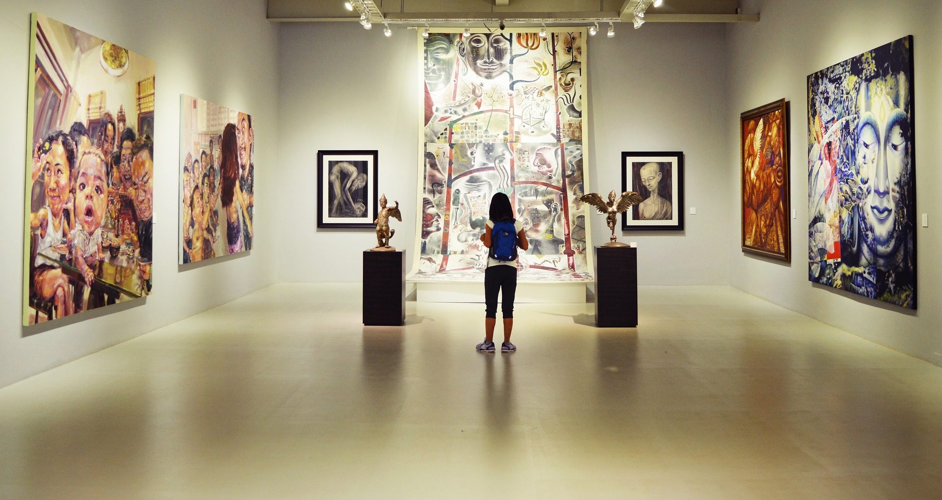 Das Foto zeigt einen Ausstellungsraum, der rechts, links und im hinteren Bereich durch drei weiße Wände begrenzt ist. Vor der hinteren Wand steht eine Besucherin mit dem Rücken zum Betrachter. Sie trägt einen Rucksack und schaut sich das Bild auf der hinteren Wand an. Das Bild reicht von der Decke bis zum Fußboden. Rechts und links des Bildes stehen zwei schwarze Sockel mit Ausstellungsobjekten. Rechts und links neben dem großen Bild hängen zwei kleinere Portraits, jeweils von schwarzen Rahmen umrahmt. An der rechten und linken Wand hängen jeweils zwei großformatige Leinwände.