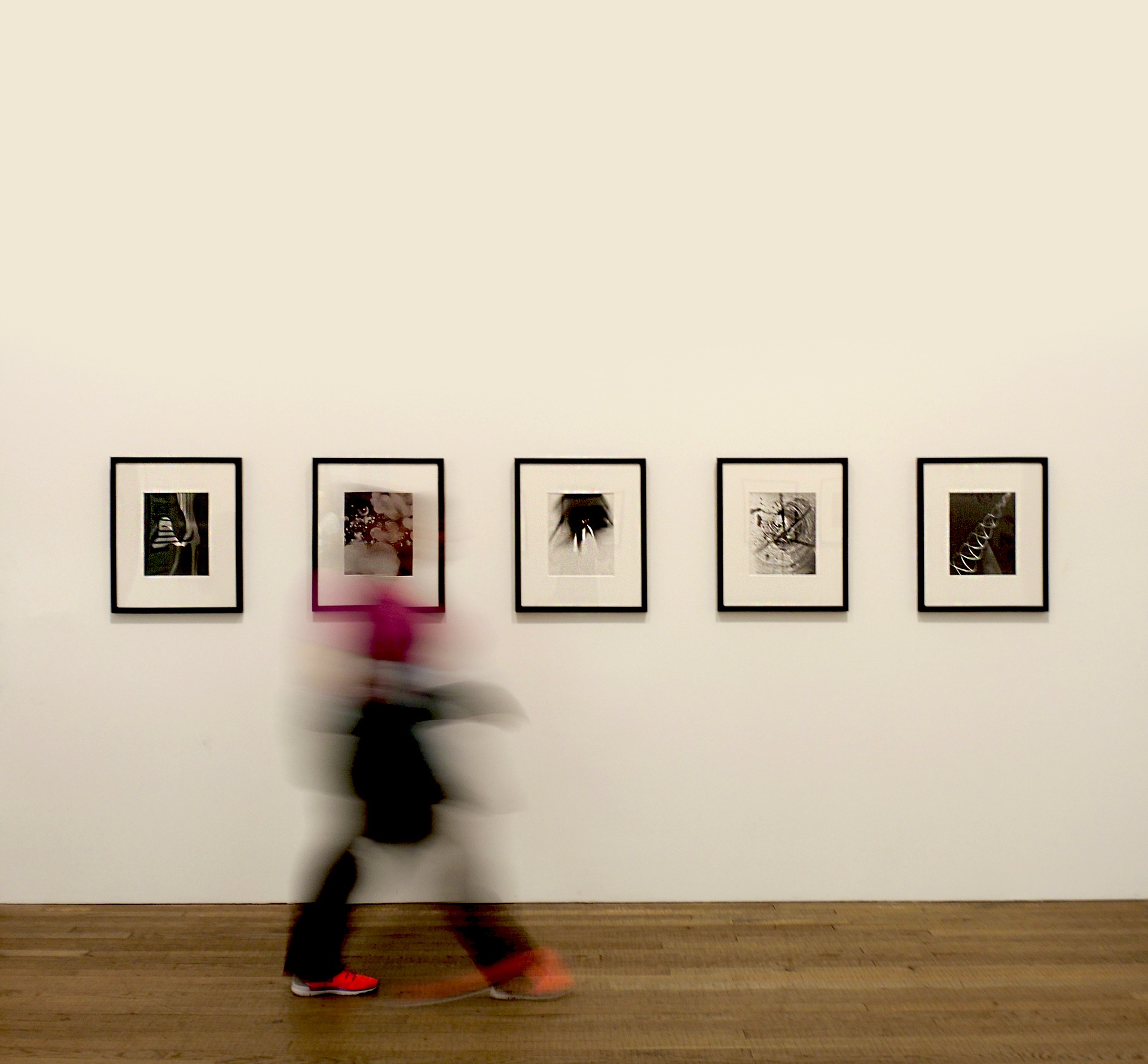 Das Foto zeigt eine weiße Wand, an der fünf schwarz gerahmte Bilder aufgehängt sind. Eine Person läuft an den Bildern von links nach rechts vorbei. Während die Bilder im Hintergrund scharf sind, ist die Person im Vordergrund verschwommen.