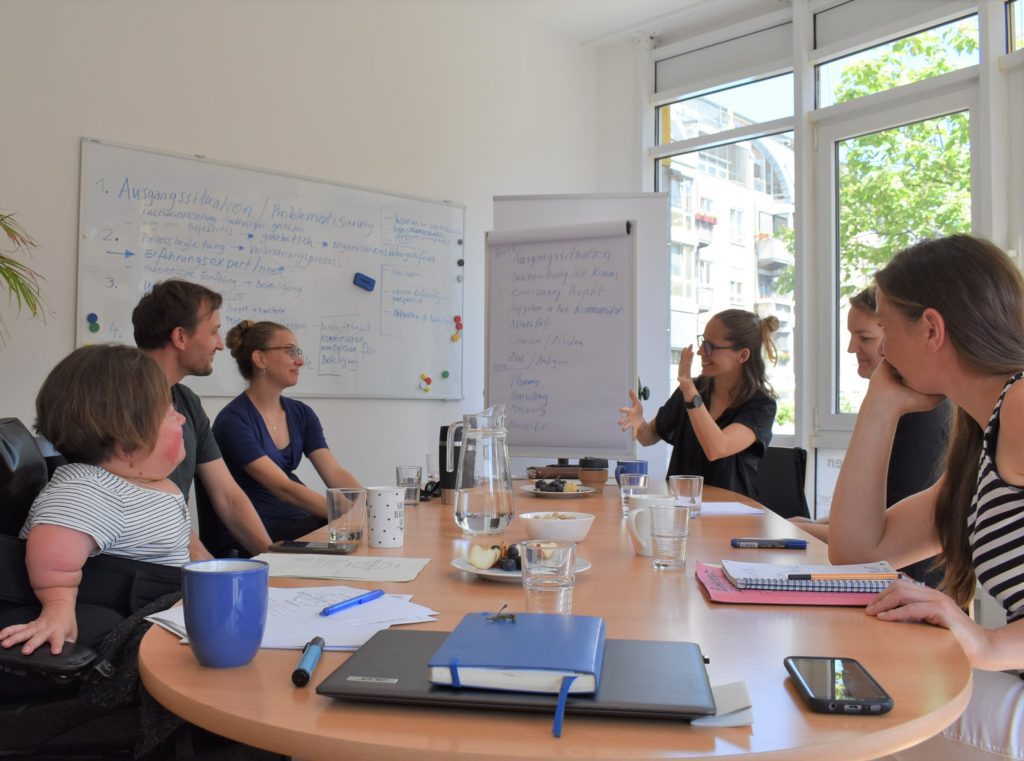 Das Foto zeigt sechs Personen, die um einen großen ovalen Tisch herum sitzen. Auf dem Tisch stehen Tassen, Gläser und Schreibzeug. Die drei Personen auf der rechten Seite schauen auf eine der gegenübersitzenden Personen, die gebärdet. Im Hintergrund sind ein beschriebenes Whiteboard und Flipchart zu sehen.