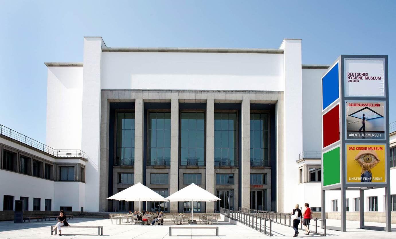 Das Bild zeigt die Fassade des Hygienemuseums in Dresden. Ein großer rechteckiger Hinterhof mit zwei weißen Sonnenschirmen. Menschen sitzen darunter. Eine lange Rampe läuft auf den Eingang des Hauptgebäudes zu. Die Fassade ist ein hoher Bau aus Beton und Glas mit einer Säulenstruktur.