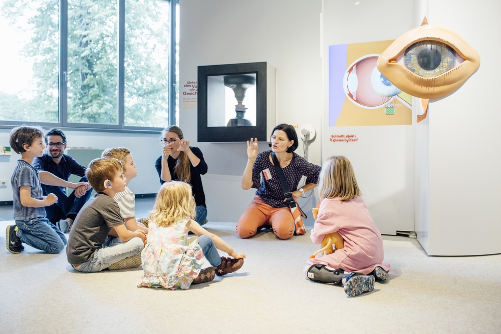 Das Foto zeigt einen Ausstellungsraum. Eine Gruppe von Kinder sitzt auf dem Boden. Zwei Frauen knien vor den Kindern und erklären etwas in Gebärdensprache. Rechts an der Wand befindet sich eine große Nachbildung eines Auges.