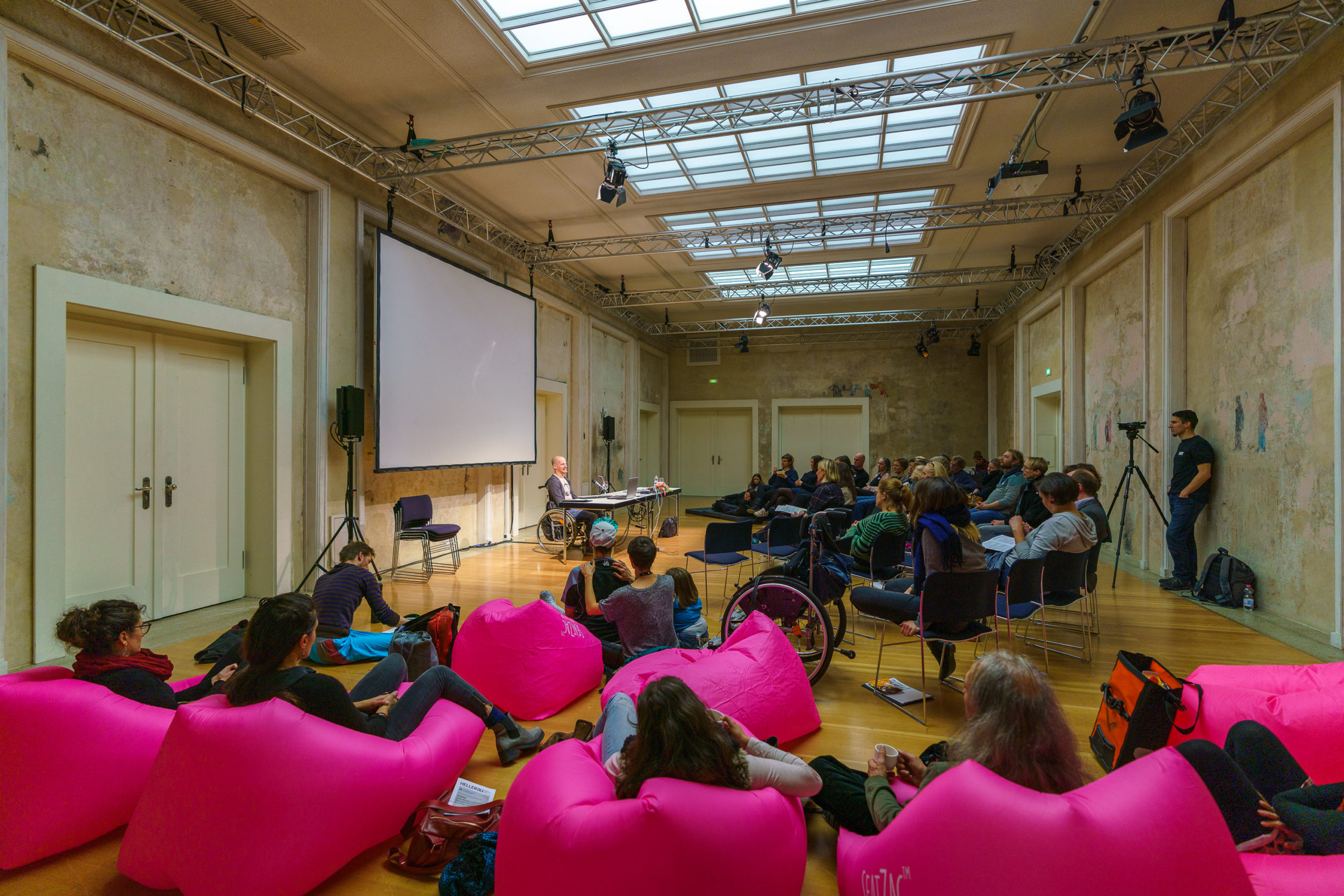 Ein länglicher Raum mit Oberlichtern. Im Vordergrund pinke Sitzsäcke mit Zuhörer:innen. Links im Raum sitzt der Referent in einem Rollstuhl an einem Tisch. Rechts sitzen weitere Zuhörer:innen, zum Teil auf Stühlen, zum Teil auf dem Boden oder auf Sitzsäcken.