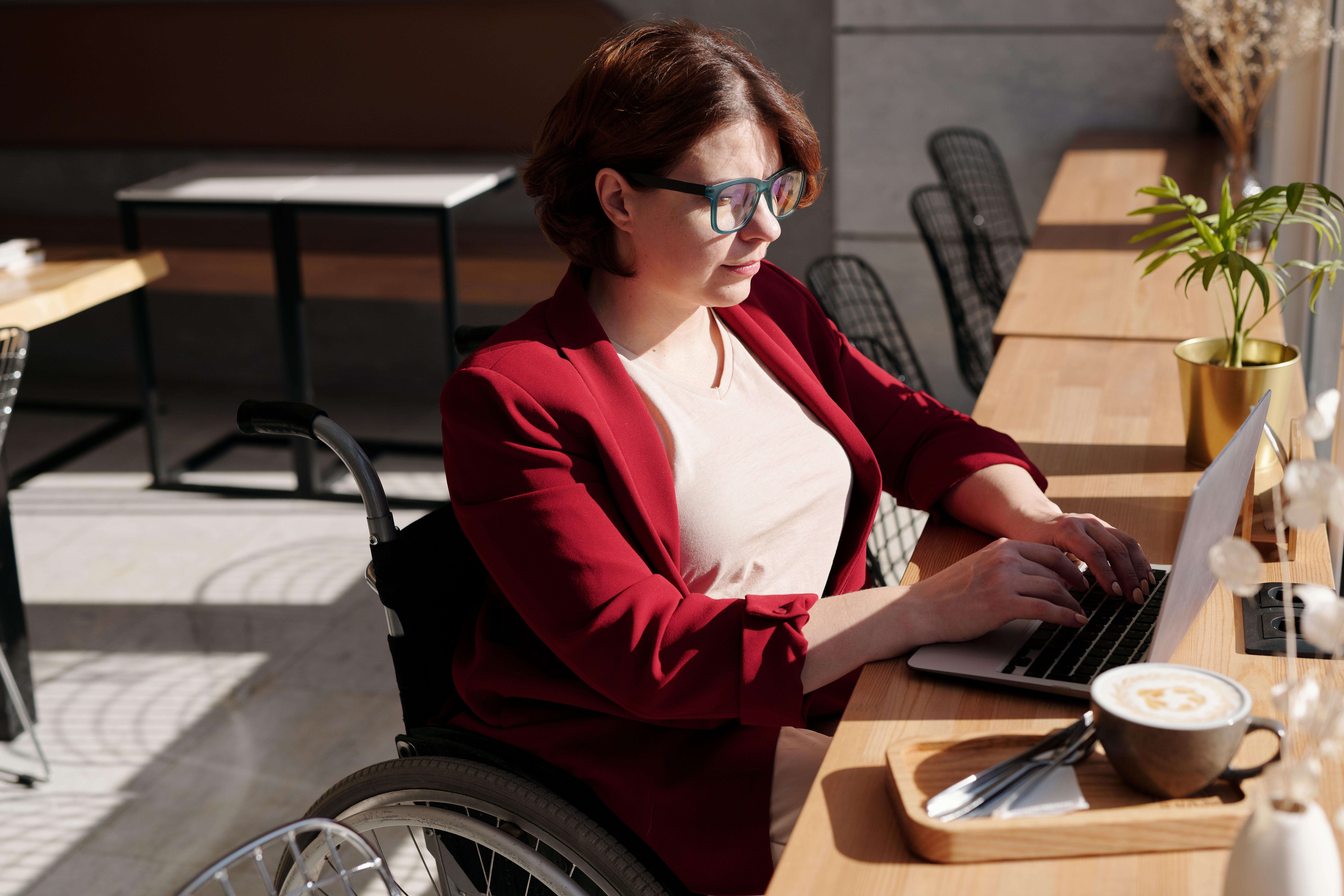 Das Foto zeigt eine Frau im Rollstuhl an einem Tisch sitzend. Vor ihr der aufgeklappte Laptop und rechts daneben eine Tasse Kaffee. Im Hintergrund stehen weitere Café-Tische.