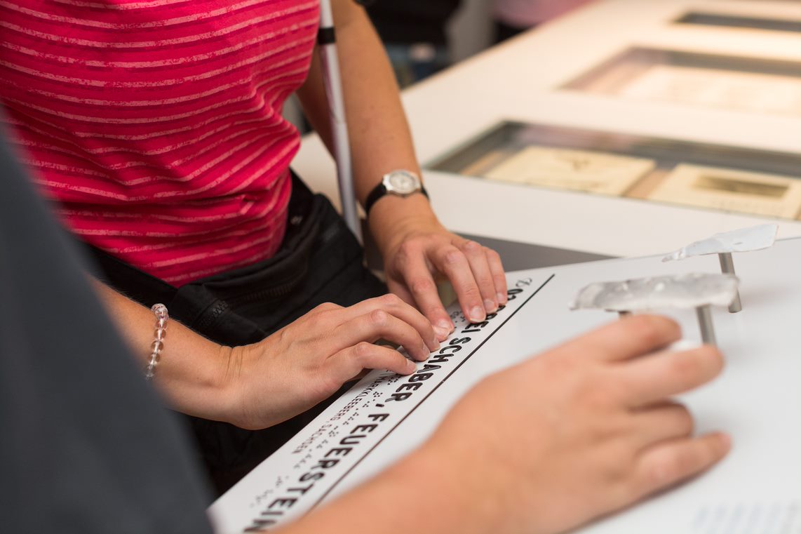 Das Foto zeigt zwei Hände, die eine Exponatbeschriftung in Relief- und Brailleschrift ertasten.