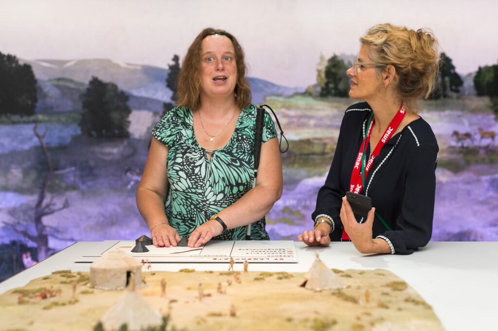 Zwei Personen stehen an einem Tisch auf dem eine historische Szenerie mit Miniaturen nachgestellt ist. Von einigen Elementen des Dioramas sind ertastbare Kopien am Tischrand platziert, die von der einen Person mit Langstock ertastet werden.