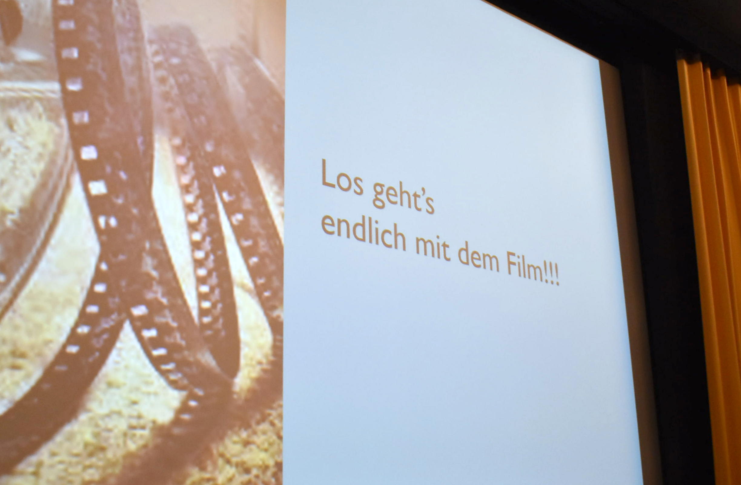 Aufnahme von schräg links unten auf Kinoleinwand. Daraul auf linker Seite alte Filmrolle und auf rechter Seite Text "Los geht's endlich mit dem Film!!!"