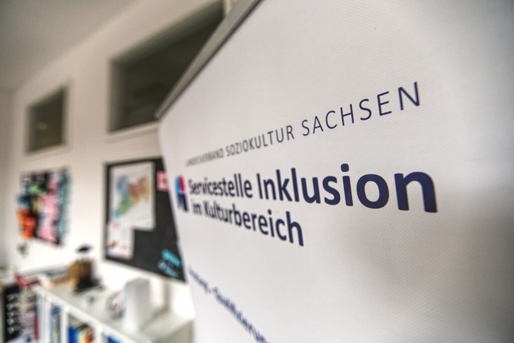 Aufnahme im Büro der Servicestelle Inklusion im Kulturbereich. Links im Vordergrund, große Teile der Bildfläche ausfüllend, oberer Abschluss eines schräg gestellten Rollups. Darauf ist das Logo der Servicestelle erkennbar.