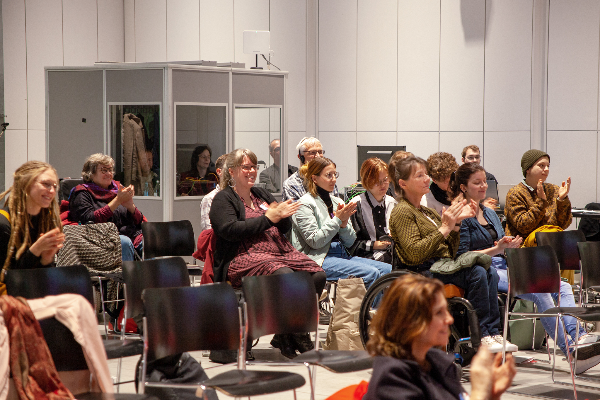 Das Foto zeigt die Konferenzteilnehmenden schräg von rechts vorn. Sie sitzen auf Stühlen und klatschen. Im Hintergrund ist eine Dolmetschkabine zu sehen.