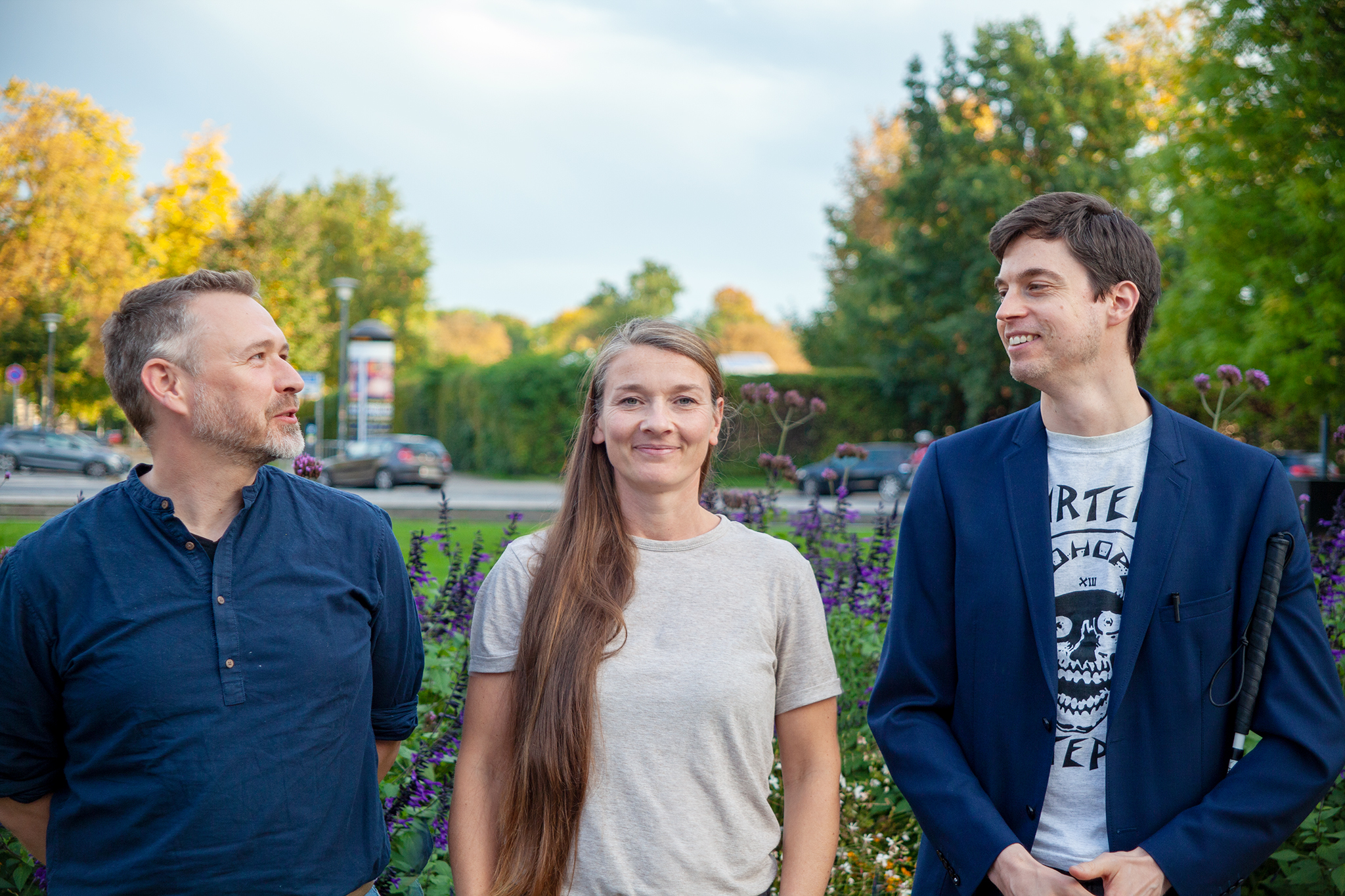 Gruppenbild der Mitarbeiter:innen der Servicestelle Inklusion im Kulturbereich (Halbportraits): In der Mitte steht Johanna von der Waydbrink frontal im hellgrauen T-Shirt, lachend mit geschlossenem Mund, die langen braunen Haare über die rechte Schulter nach vorn und die Arme an die Seite gelegt. Links steht Matthias Franke im kragenlosen dunkelblauem Hemd, die Arme nach hinten genommen. Mit kurzen graumelierten Haaren und Bart blickt er scherzend zum rechts stehenden Dirk Sorge. Dieser mit kurzen dunkelbraunen Haaren, im dunkelblauem Jackett, darunter ein hellgraues T-Shirt und einen Langstock an seiner linken Seite, blickt lächelnd zu M. Franke. Im Hintergrund befindet sich ein Blumenbeet und eine Straße.