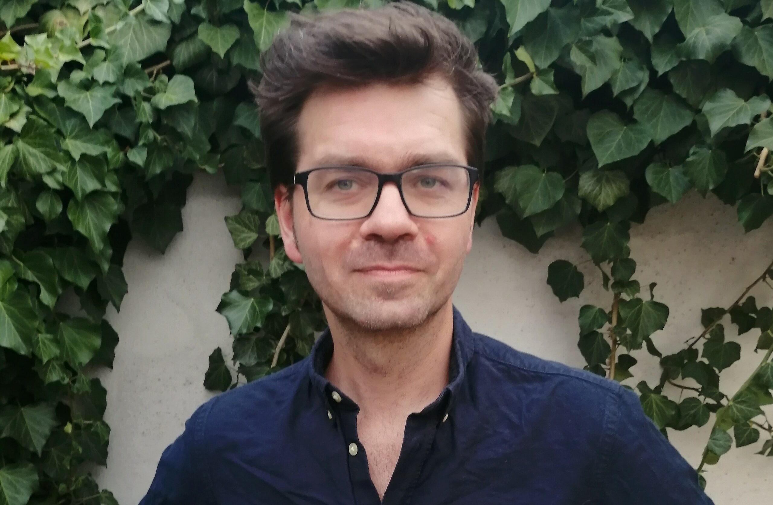 Das Foto zeigt Sebastian Göschel. Er hat dunkelbraunes Haar, trägt eine Brille mit schwarzem Gestell und ein dunkelblaues Hemd. Im Hintergrund eine mit Efeu bewachsene Wand.