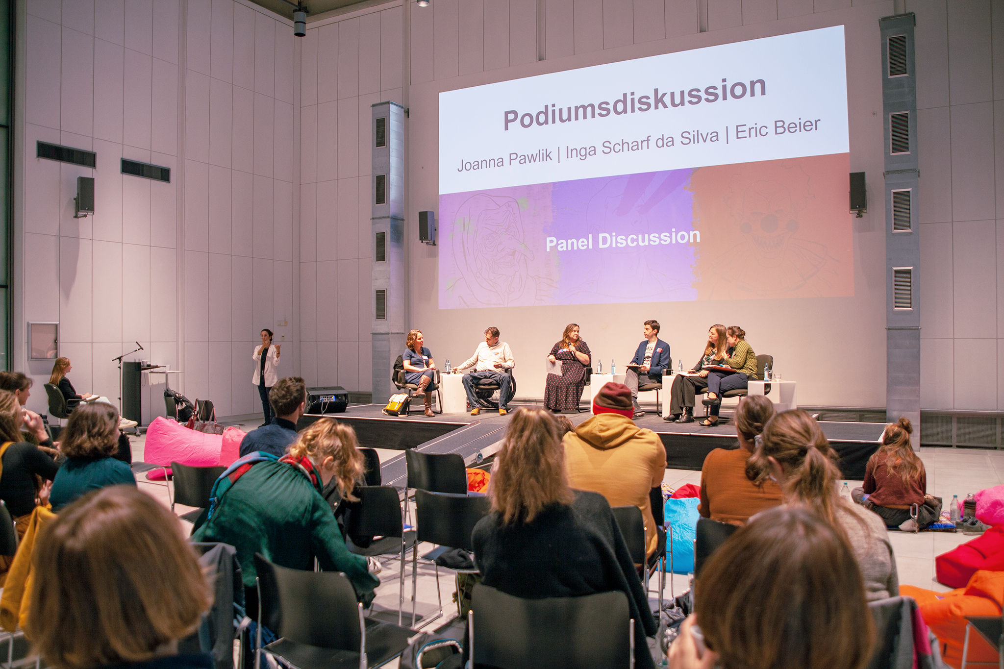 Das Foto zeigt das Konferenzpodium im Hintergrund und die Teilnehmenden, die auf das Podium schauen, von hinten, im Vordergrund. Auf dem Podium sitzen sechs Personen. Hinter den Podiumsgästen ist eine Projektion mit der Beschriftung "Podiumsdiskussion" zu sehen.