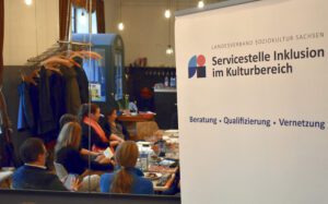 Im Hintergrund sitzen Menschen an einem Tisch. Im Vordergrund steht ein Aufsteller der Servicestelle Inklusion im Kulturbereich.