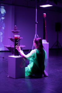 Das Bild zeigt eine Künstlerin von hinten, hockend. Sie greift mit ihrem linken Arm an einen gläsernen Zylinder in dem mutmaßlich Bakterienkulturen angesiedelt sind. Die Bühne ist in warmes, violettes Licht getaucht.