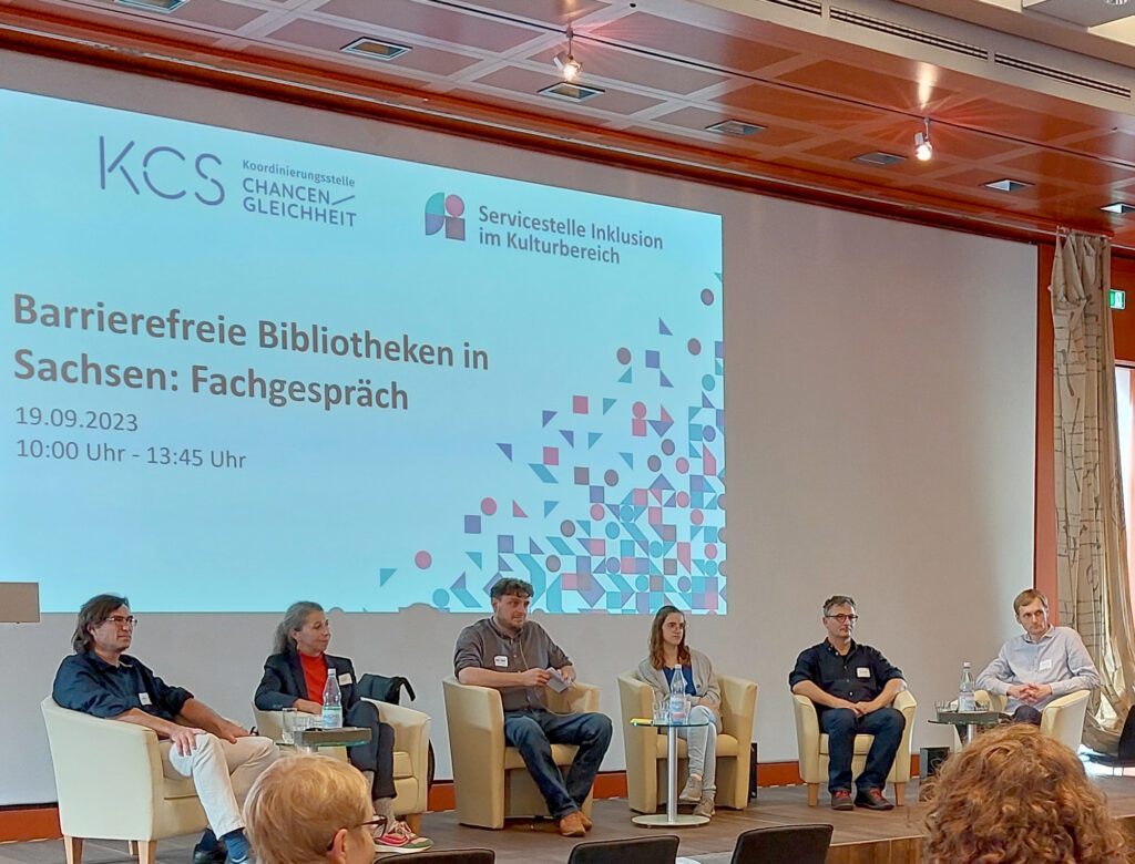 Blick auf das Podium während der Fachveranstaltung. Auf dem Pudium sitzen die Referent:innen und der Moderator. Im Hintergrund Baemerprojektion mit Veranstaltungsankündigung. Vor angeschnitten, Köpfe der Teilnehmer:innen.