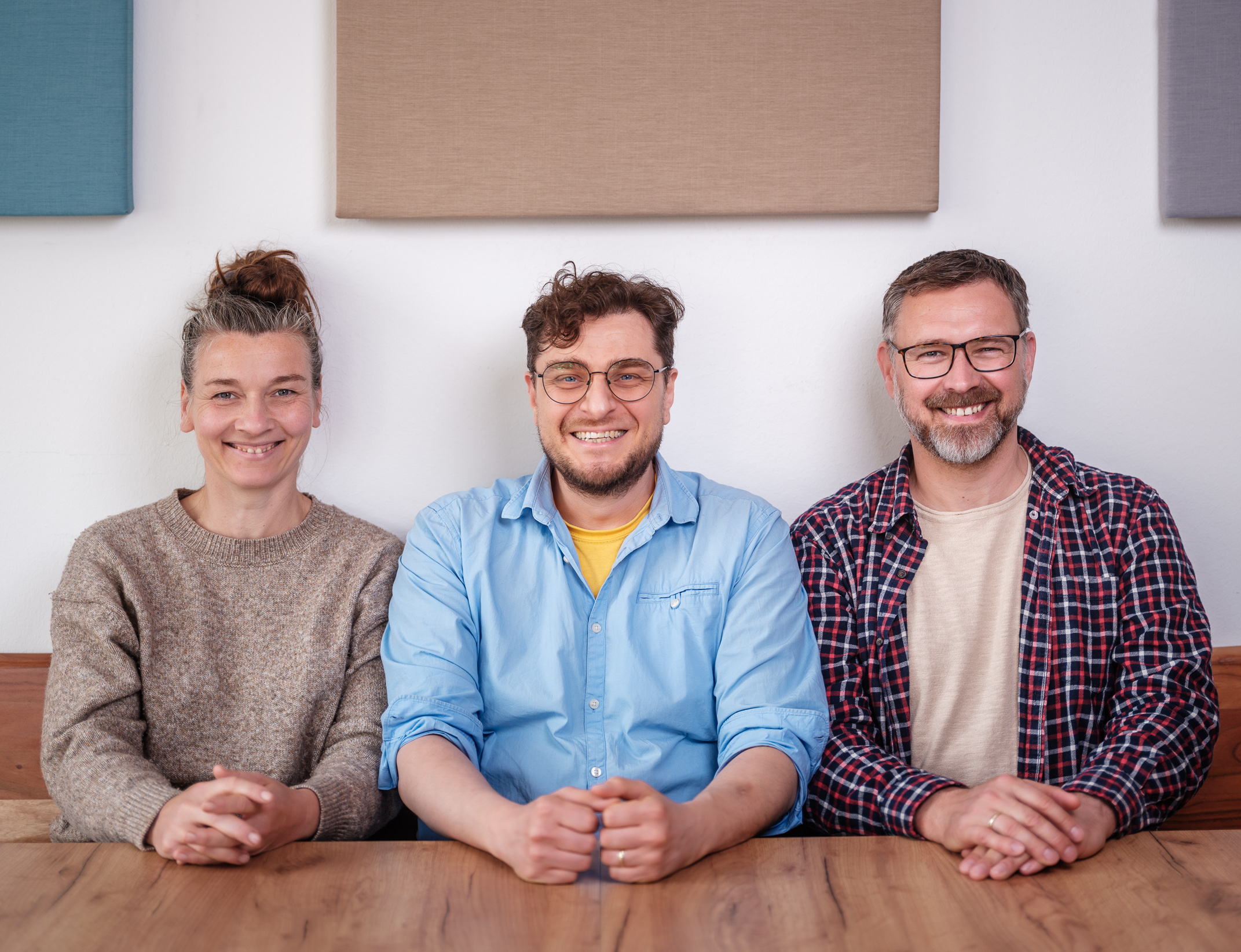 Auf dem Bild sitzen von links nach rechts Johanna von der Waydbrink, Eric Beier, Matthias Franke nebeneinander an einem Holztisch. Johanna von der Waydbrink trägt einen beigefarbenen Strickpullover und hat die Hände überkreuzt, Eric Beier trägt ein hellblaues Hemd über einem gelben T-Shirt und hat die Hände vor sich auf dem Tisch gefaltet, Matthias Franke ist mit einem karierten Hemd über einem beigen T-Shirt bekleidet und hält ebenfalls die Hände gefaltet vor sich. Der Hintergrund zeigt eine weiße Wand mit rechteckigen, schallabsorbierenden Paneelen in verschiedenen Farben.