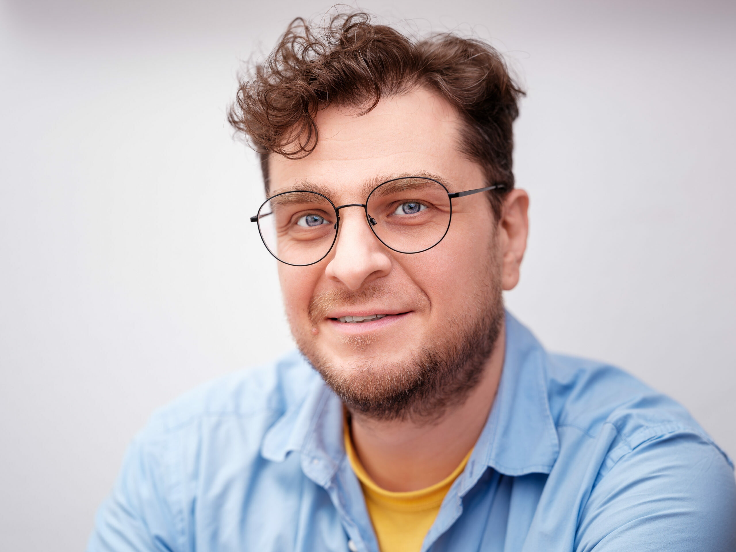 Eric Beier mit lockigen braunen Haaren, Brille und kurzem Bart trägt ein hellblaues Hemd über einem gelben T-Shirt. Der Hintergrund ist neutral und hell gehalten.