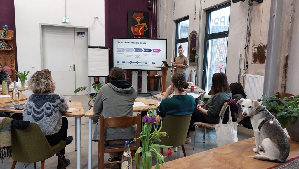 Das Foto zeigt eine Sitzung in einem offenen, modern eingerichteten Raum. Mehrere Personen sitzen an Holztischen und hören Johanna von der Waydbrink zu, die vorne an einem Stehpult steht und eine Präsentation hält. Auf dem großen Bildschirm hinter ihr steht die Überschrift „Phasen der Prozessbegleitung“ mit einem farbigen Ablaufdiagramm. Im Vordergrund stehen Blumenvasen auf den Tischen, und am rechten Bildrand sitzt ein weiß-grauer Hund auf einem erhöhten Podest und schaut in die Kamera. Die Atmosphäre wirkt konzentriert und entspannt.